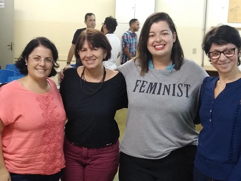 Quatro mulheres, em pé, sorrindo, abraçadas. Professora Camila de cabelo médio preto, óculos, pele clara, blusa rosa. Professora Flomar, cabelo curto, castanho, pele clara, blusa preta e colar escuro. Mestranda Karine, cabelo castanho nos ombros, liso, pele clara, camiseta cinza escrito Feminist. Professora Vanderleida, pele clara, cabelo curto e preto, óculos, bata azul marinho