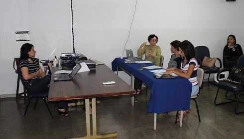 As professoras estão sentadas à mesa da banca, de frente para a aluna, também sentada em sua mesa. A professora Joana está falando e olhando para a professora Marlei.