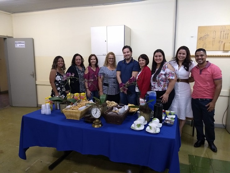 Na sala de defesa do Programa, de pé, atrás de uma mesa decorada com flores, com forro azul e com um rico café da manhã, encontram-se, respectivamente da esquerda para a direita, Tattiana (egressa), Luciene (professora), Marta (professora), Elismar (egressa), Ulysses (egresso), Flomar (professora), Caroline (egressa), Lílian (egressa), Thiago (egresso). Todos estão vestidos informalmente. 